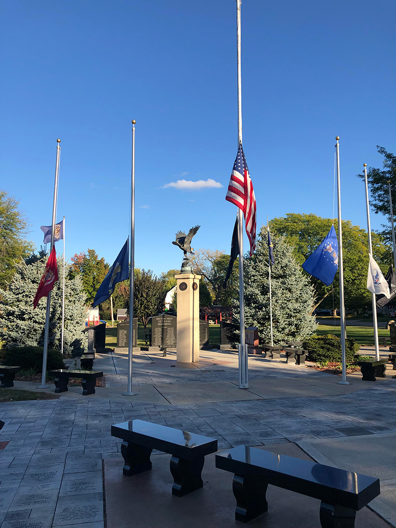Flagpoles, trees, stone memorials, and benches under a clear blue sky